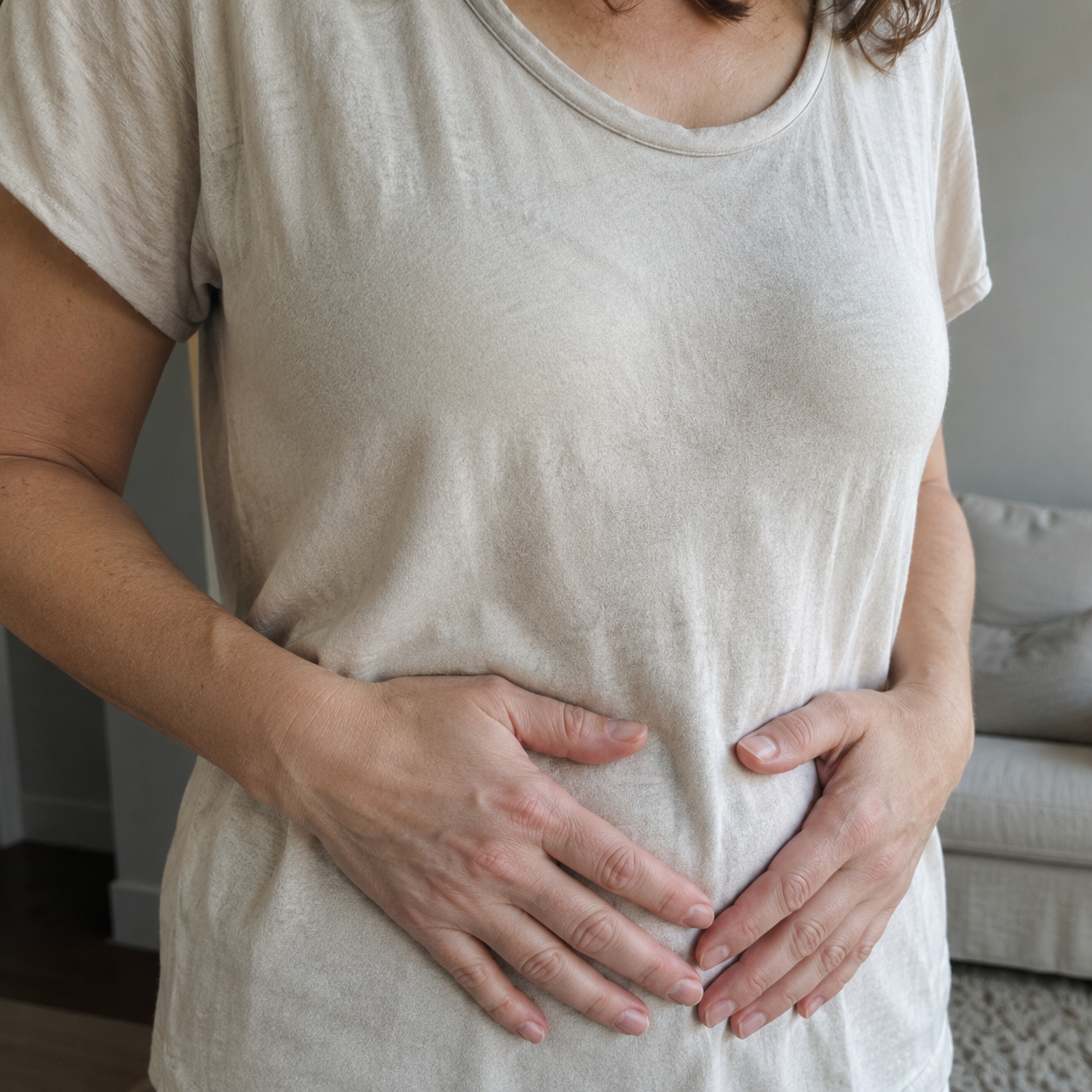 A woman's torso from chest to hip, both hands resting gently on her lower stomach, wearing a soft cream cotton tee.